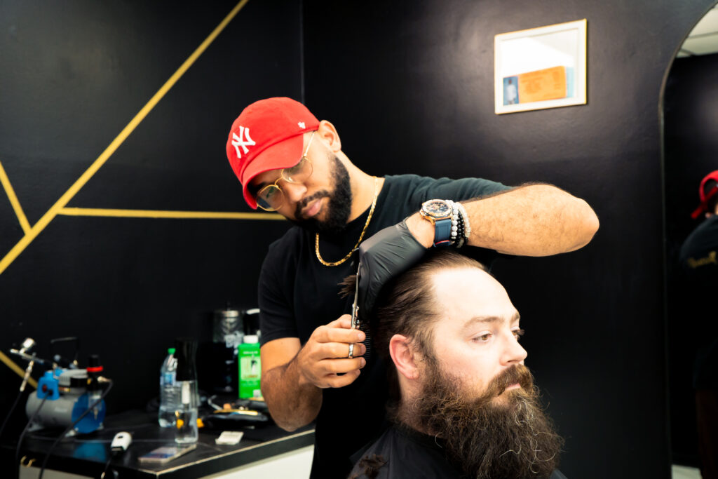 A Golden Barbershop barber cuts a bearded client's hair in the shop's modern interior with dark walls and gold accents.