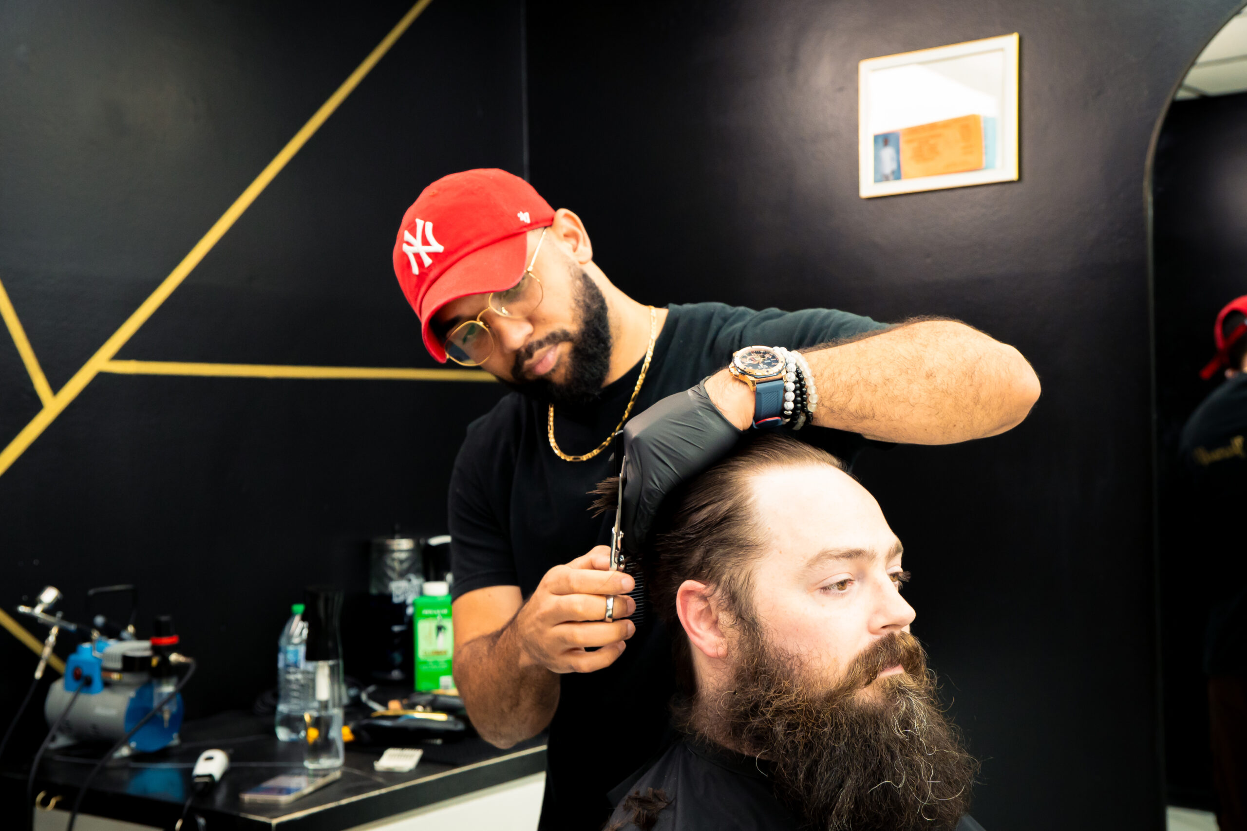 A Golden Barbershop barber cuts a bearded client's hair in the shop's modern interior with dark walls and gold accents.
